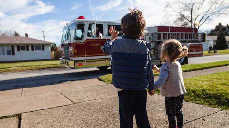 Two young children wave at a passing fire truck from their driveway.